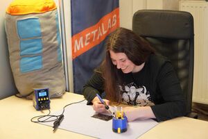 Eine Person mit langen braunen Haaren sitzt an einem Tisch und lötet lächelnd an einer Platine. Auf dem Tisch stehen ein blauer Lötkolben, eine Lötstation mit Temperaturanzeige (370°C) und ein Werkzeughalter mit weiteren Lötwerkzeugen. Im Hintergrund hängt eine Fahne mit der Aufschrift „METALAB“ sowie ein großes Stoffkissen in Form einer Telefonzelle. Die Person trägt ein schwarzes T-Shirt mit einem weißen Tiermotiv.